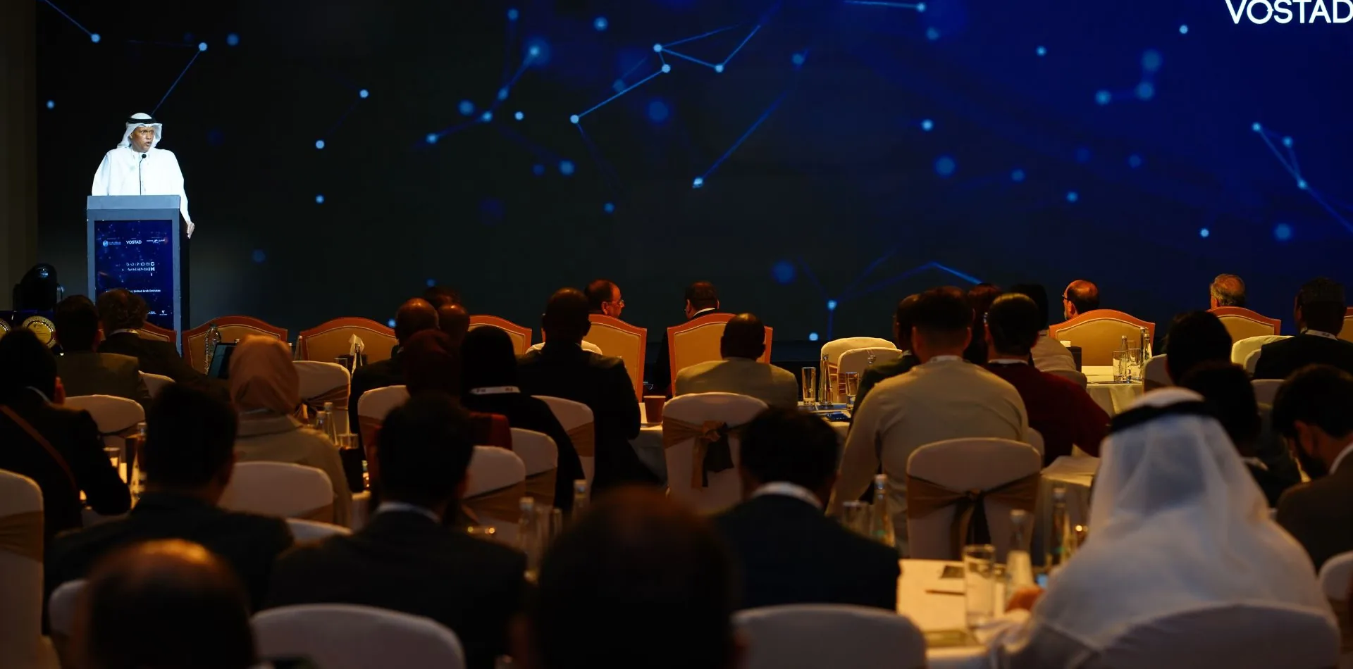 A speaker at the podium before a full room of delegates at round tables